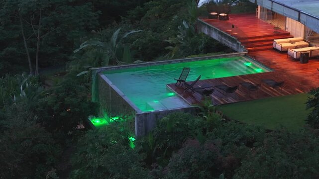Aerial twilight view of modern luxury villa with illuminated infinity pool featuring color-changing LED lights, wooden deck terrace with lounge furniture, surrounded by lush tropical jungle foliage.