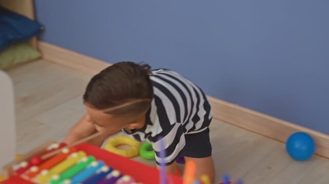 Little boy reaches with hand to grab colorful toy rings on floor inside building; playful curiosity.