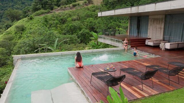 Group of young people dancing by pool at small party surrounded by lush jungle hills. Man dives into turquoise infinity pool while friends swim and relax on wooden deck with cocktails at modern villa.