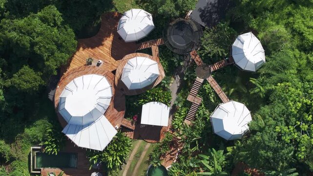 Aerial drone footage captures a luxury glamping resort featuring white geodesic dome tents connected by wooden decks and walkways, nestled within dense tropical rainforest canopy on a sunny day.