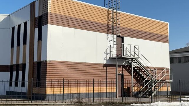 Stylish modern house with siding under blue sky, geometric facade of glass and wood panels, parking lot and metal fence