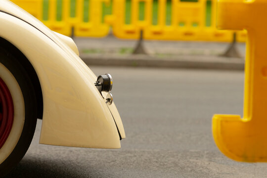 Tight framing of a cream-colored classic car front fender and round headlight parked near a yellow plastic road barrier on asphalt. The scene shows painted