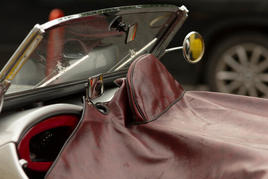 Close-up view of a classic convertible cockpit featuring a folded windshield, chrome side mirror, and a worn burgundy leather tonneau cover draped over the
