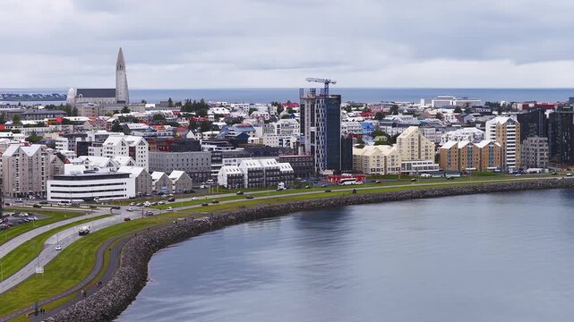 Aerial daytime view of Reykjavik, Iceland, shows Hallgrimskirkja, curved seawall, ring road, traffic, water ripples, new high rises, crane, and overcast sky.