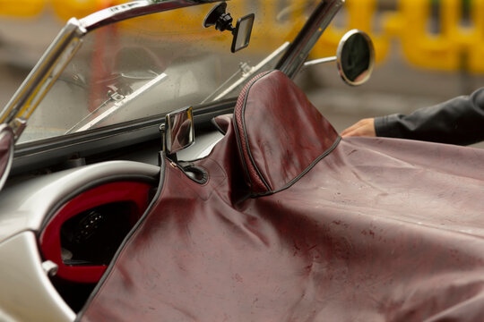 Tight view of a vintage convertible's cockpit featuring a burgundy leather tonneau cover stretched over the passenger area, a small round side mirror, chrome