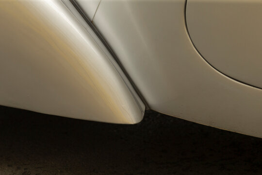 Detailed close-up showing a light-colored car body panel and adjacent wheel arch with smooth painted metal surfaces, subtle reflections, and a dark textured