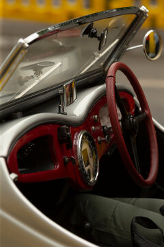Close-up view of a vintage sports car interior featuring a textured red steering wheel, polished chrome dashboard trim, circular gauge cluster, and small