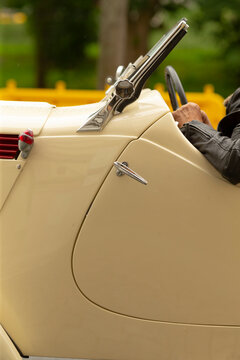 Tight view of a cream-colored vintage convertible showing the curved door, chrome door handle and hood ornament, and partial driver wearing a dark jacket