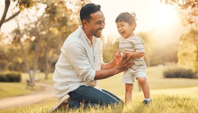 A dark-haired man kneeling on grass holding a toddler's hands in a park with warm sunlight and trees in the background.