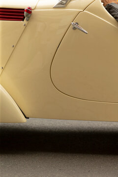 Close-up view of the side panel of a cream-colored vintage car showing smooth painted metal, a small chrome door handle, curved fender edge and a red louvered