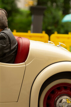 Partial side view of a vintage cream convertible showing the rear wheel, fender, and red leather passenger seat. A person wearing a dark jacket sits in the