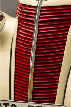 Tight detail of a classic automobile front showing a vertical chrome center trim and a red slatted grille set into a cream painted body. Smooth glossy paint