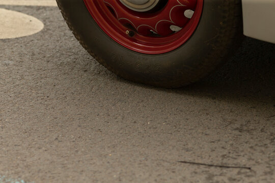 Low-angle close view of a red steel wheel and black tire of a vintage car standing on textured asphalt. The scene shows tire tread, painted steel rim