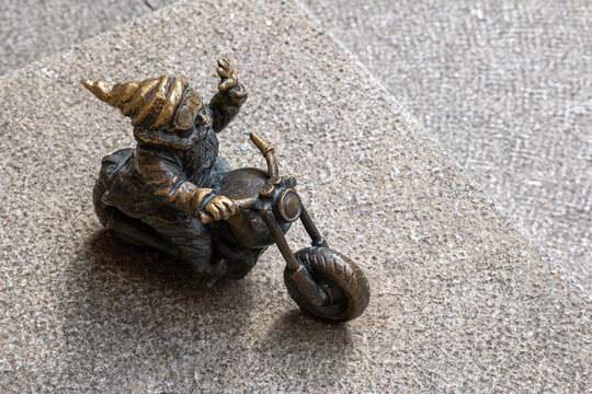 Small bronze Wroclaw dwarf statue riding motorcycle on granite pavement, closeup of whimsical street art, tourist landmark, urban folklore, travel souvenir, public sculpture, local culture, no people