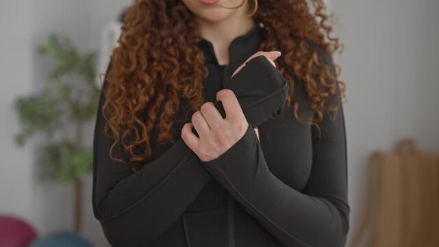 Young curly woman holding wrist and adjusting sleeve before yoga session in studio; mindful calm recovery.