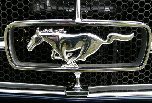 Close-up of a chrome Ford Mustang running horse emblem mounted on a honeycomb grille, highlighting automotive design, vintage styling, metallic textures and motion-inspired branding on a classic car.