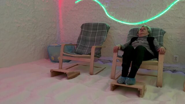 Elderly woman reclining in salt chamber, wearing cap and seated on wooden chair with footstool, legs extended over salt crystal