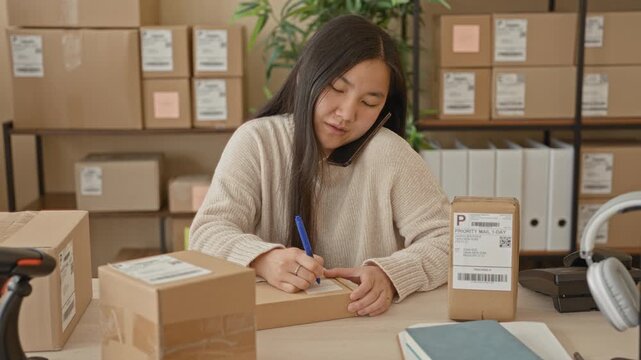 Woman writing an address on a parcel with pen and hand while speaking on smartphone in a small business building; productivity.