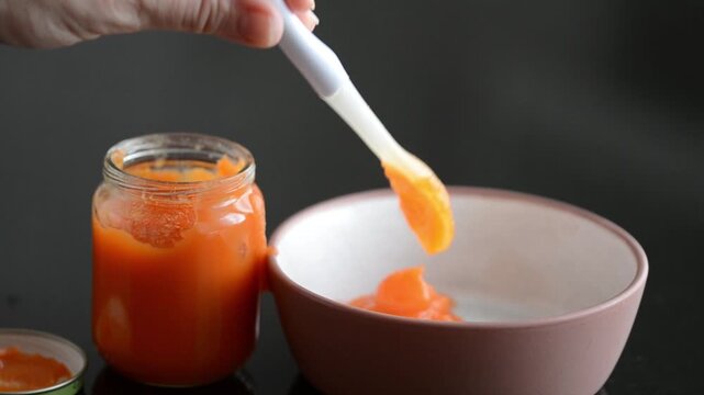 Baby food carrot puree being poured from a glass jar onto a plate on a black background