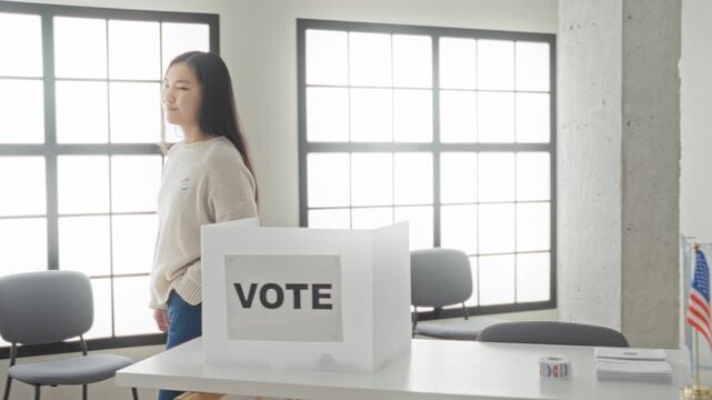 Young chinese woman beside white vote box, hand showing two fingers and wearing sticker at voting booth in building;civic pride.