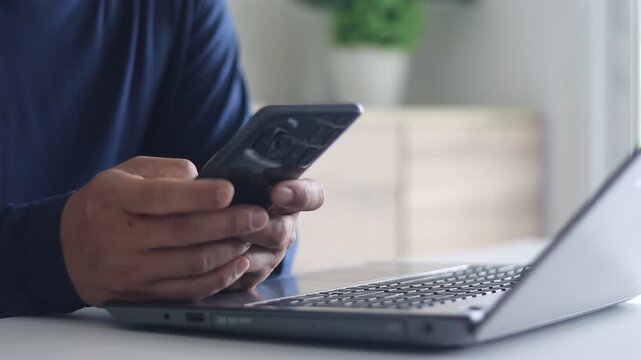 Businessman hand typing mobile phone while working on  laptop. Man chatting online using a smartphone. Male hands scrolling, typing a message on a phone. Business technology communication.