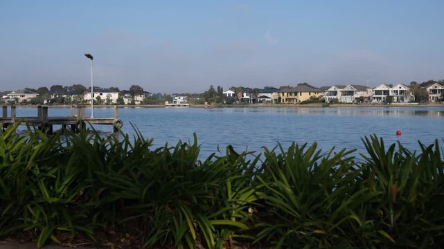 Waterfront homes or upscale houses surround a tranquil lake at Sanctuary Lakes, Point Cook, viewed past a jetty. Premium lakeside living in Melbourne's western suburbs in Australia