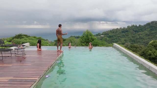 Group of young people dancing by pool at small party in tropical resort. Tattooed man stands on wooden deck while women swim and socialize in turquoise infinity pool overlooking lush jungle and ocean.