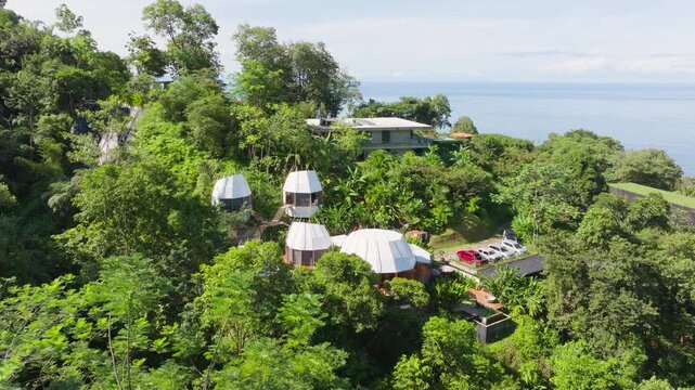 Aerial view of exclusive villa in Costa Rica featuring unique dome-shaped bungalows surrounded by dense tropical rainforest on hillside overlooking Pacific Ocean with parked cars and modern.