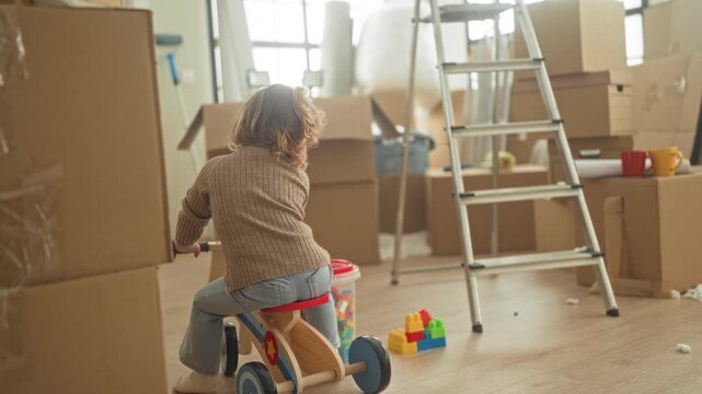 Girl riding a wooden ride on toy past stacked moving boxes and aluminum ladder inside a building, looking back while playing with colorful blocks; playful curiosity.