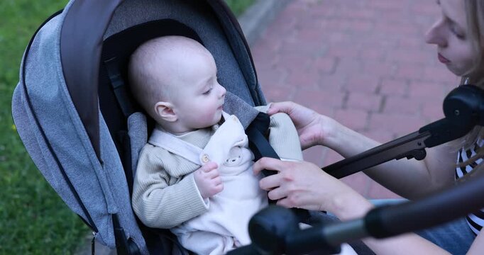 Mother buckling up her cute baby in stroller at park. Camera moving forward