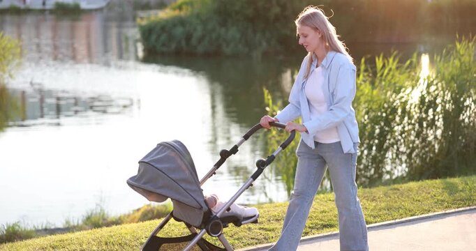 Mother walking with her cute baby in stroller at park on sunny day. Camera moving left