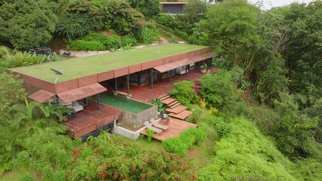 Aerial drone footage captures stunning modern tropical villa with green grass roof, wooden decks, infinity pool and lounging areas nestled in dense Costa Rican rainforest hillside on overcast day.