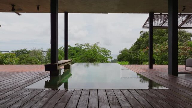 Stunning villa in Costa Rica features an infinity pool with wooden deck surrounded by lush tropical rainforest. Modern architecture with dark pillars frames the serene jungle view under overcast.