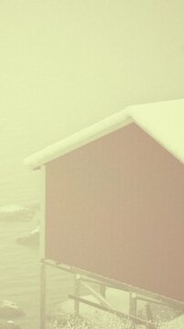 A red cabin stands near the water with snow on its roof. Thick fog surrounds the area, making visibility low. The scene captures winter weather and its effects on the landscape.