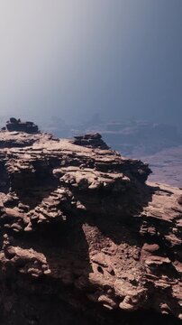 A rocky terrain shows layered stone formations under bright daylight. The view captures distant mountains and a clear sky above, highlighting the natural structure.