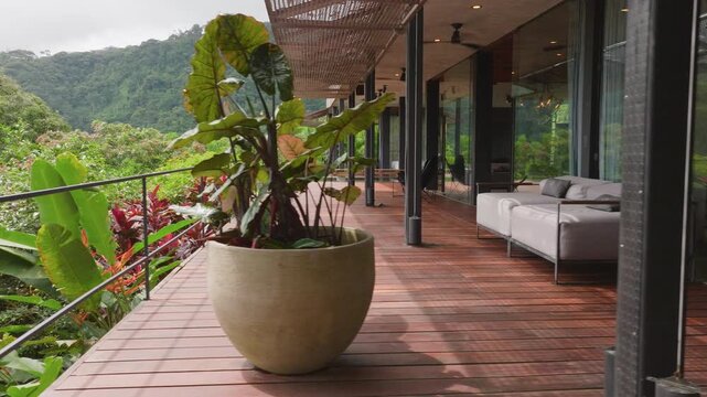 Contemporary glass and steel villa with infinity pool sits on lush tropical hillside surrounded by dense green rainforest vegetation, featuring wooden deck terraces, modern outdoor furniture.