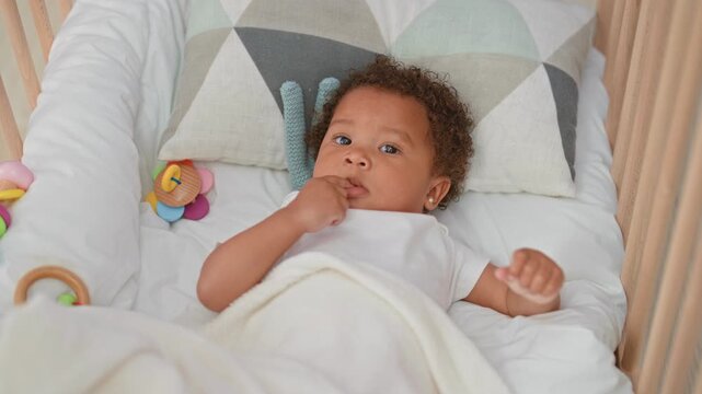 Little girl reaching for a colorful rattle in a crib nursery with blanket and pillow; curiosity bonding.