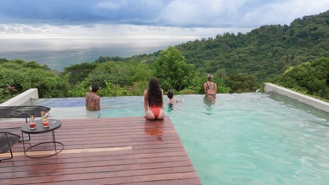 Group of young people dancing by pool at small party in luxury resort. Friends enjoy cocktails on wooden deck with infinity pool overlooking lush green rainforest mountains and ocean in slow motion.