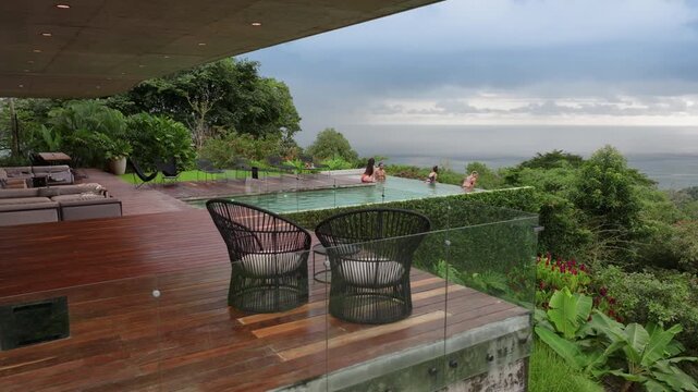Group of young people enjoying small party by luxury infinity pool overlooking tropical jungle and ocean. Friends in swimwear relax on wooden deck with modern lounge furniture under cloudy sky.