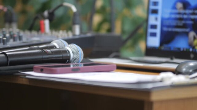 Professional audio setup on wooden desk. Two microphones sit near pink phone. Laptop displays live event. Background shows greenery. Ideal for conferences, webinars, or live streaming