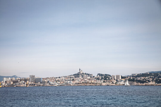 Panorama of Notre Dame de la Garde, also known as Bonne Mere, rising above the city skyline, waterfront buildings, sailboat traffic and Mediterranean coast in Marseille, France.