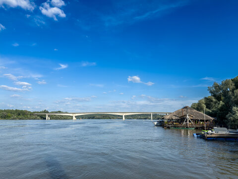 Panoramic view of the Sava River at Izletiste Zabran, in Obrenovac, Serbia, with Most na Savi bridge in the background, riverside leisure area, floating dock, splavovi, trees and open sky.