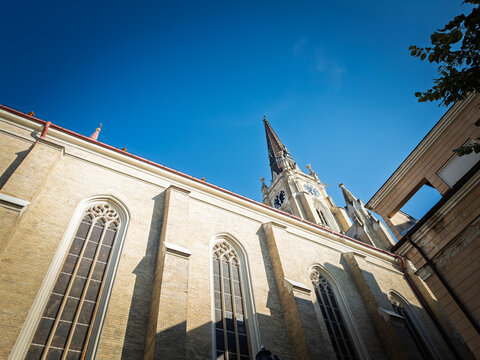 Low angle view of the Name of Mary Church, often called Novi Sad Cathedral, showing Gothic Revival windows, brick facade, clock tower and patterned spire under a blue sky in Novi Sad, Serbia.