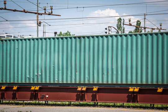 Green shipping container carried on a rail freight wagon, with overhead electric lines, industrial track equipment and a cropped view emphasizing multimodal cargo transport, logistics infrastructure.