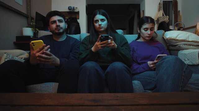 Group of young Hispanic friends sitting on a sofa ignoring a movie on TV while using their smartphones, highlighting digital distraction, divided attention, and reduced social interaction at home.
