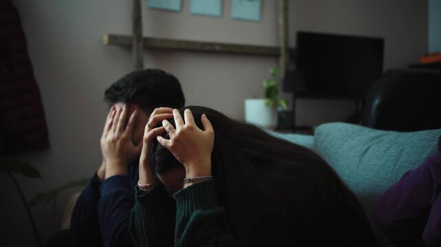 Two young Hispanic women and one young Hispanic man sitting on a sofa watching a sports game on TV, reacting with nervous anticipation before celebrating, only to be disappointed when the team misses 