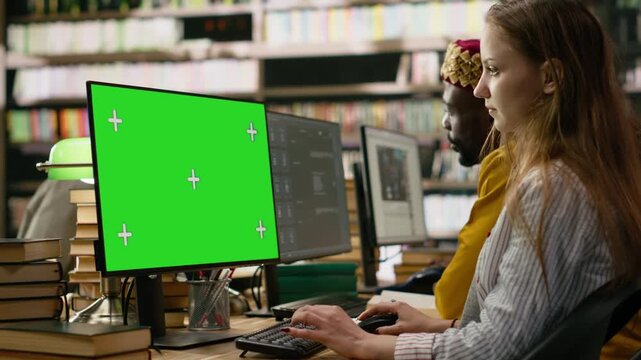 Isolated green screen on desktop monitor with tracking markers on wooden library study desk, peers studying beside books, keyboards and bookshelves, computer class, for education technology.