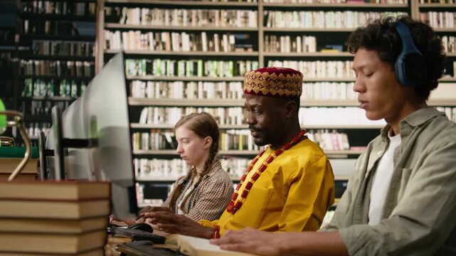 Adult student in traditional clothing work at desktop along shared library table with books, keyboards and monitors, for digital learning and academic training.