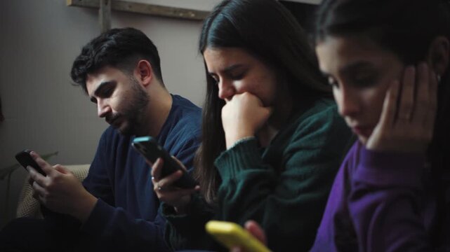 Two young Hispanic women and one young Hispanic man sitting together while watching TV, distracted by their smartphones instead of paying attention to the movie, highlighting digital distraction and r