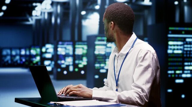 African American computer scientist writes server script code on a laptop terminal in a high tech facility, supporting software updates, maintenance work and data center operations.